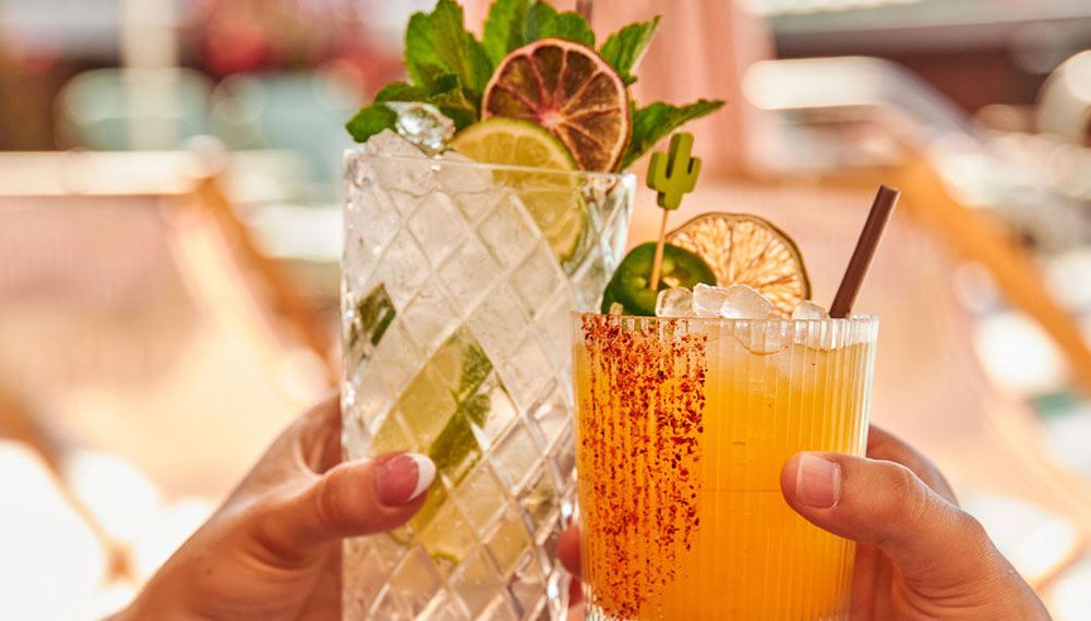 Two guests toast with handcrafted citrus cocktails garnished with lime, mint, and dried citrus slices while relaxing beside a sunlit pool lounge area.